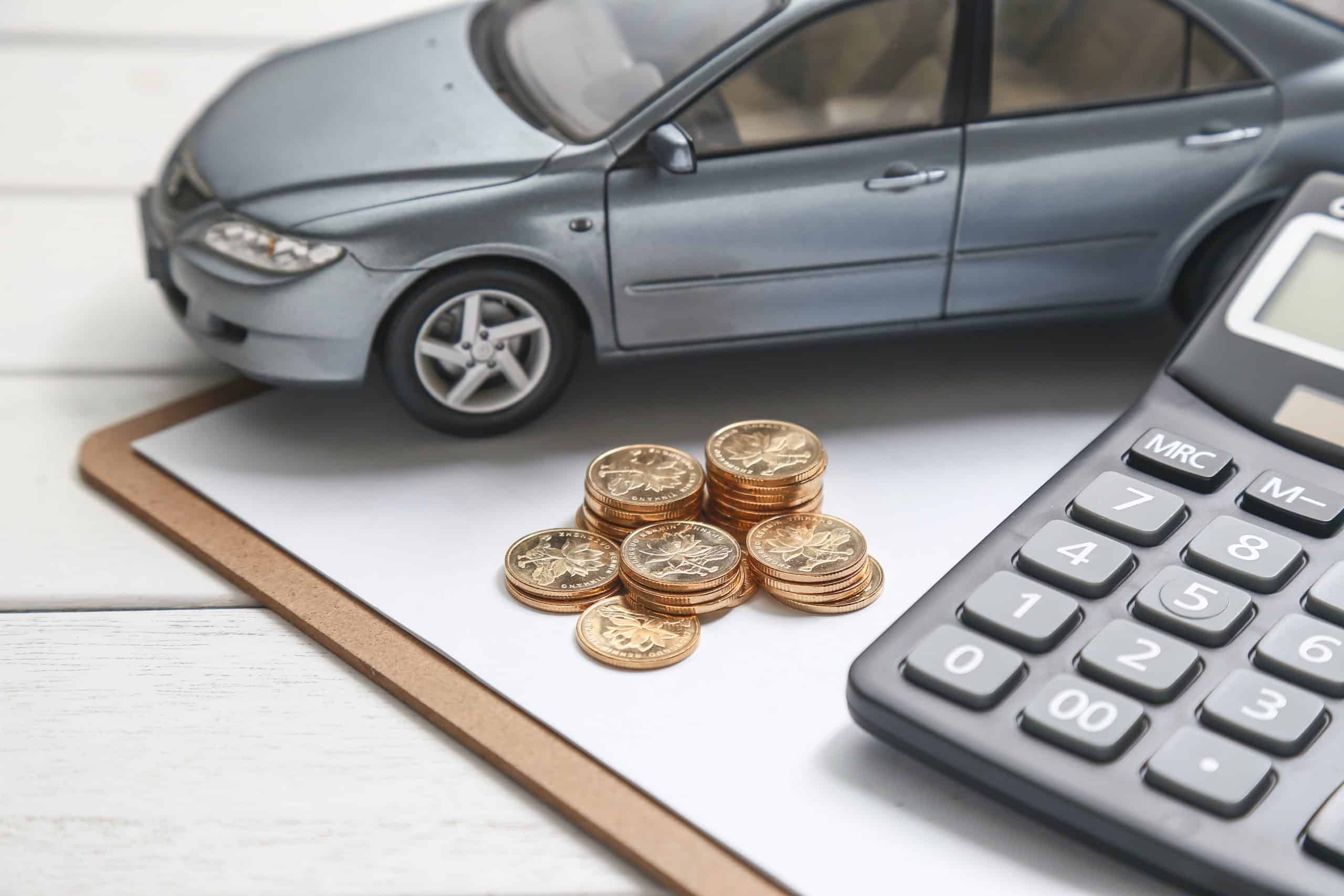 A small model of a silver car is placed on a clipboard alongside stacks of gold coins and a calculator on a white wooden table. The image symbolizes financial planning for car purchases, budgeting, or loan calculations, highlighting the importance of cost management when acquiring a vehicle.