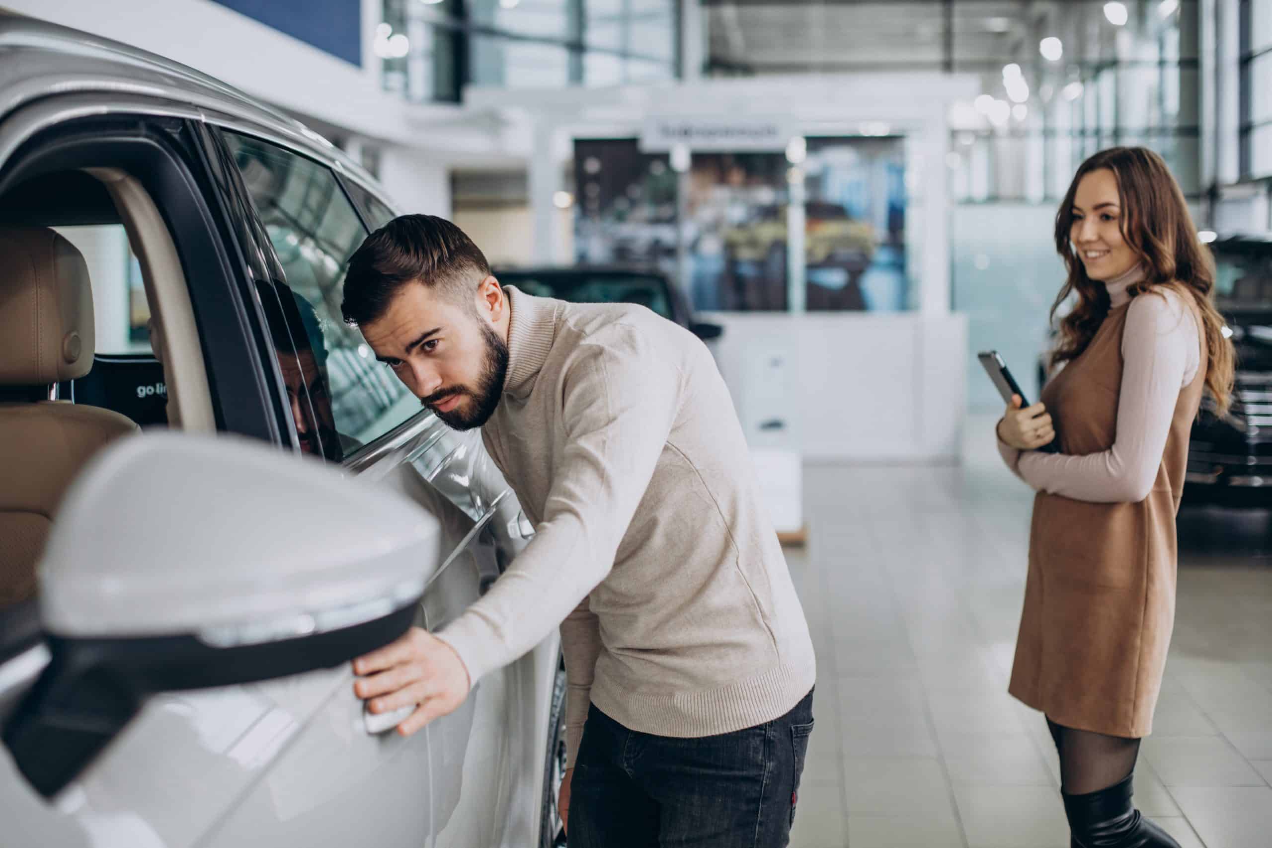 A man carefully inspects a silver car inside a modern showroom, examining its exterior with a focused expression. He appears to be considering a purchase, while a smiling female sales representative stands nearby holding a tablet, ready to assist. The bright and spacious dealership environment suggests a professional and welcoming atmosphere for potential buyers.