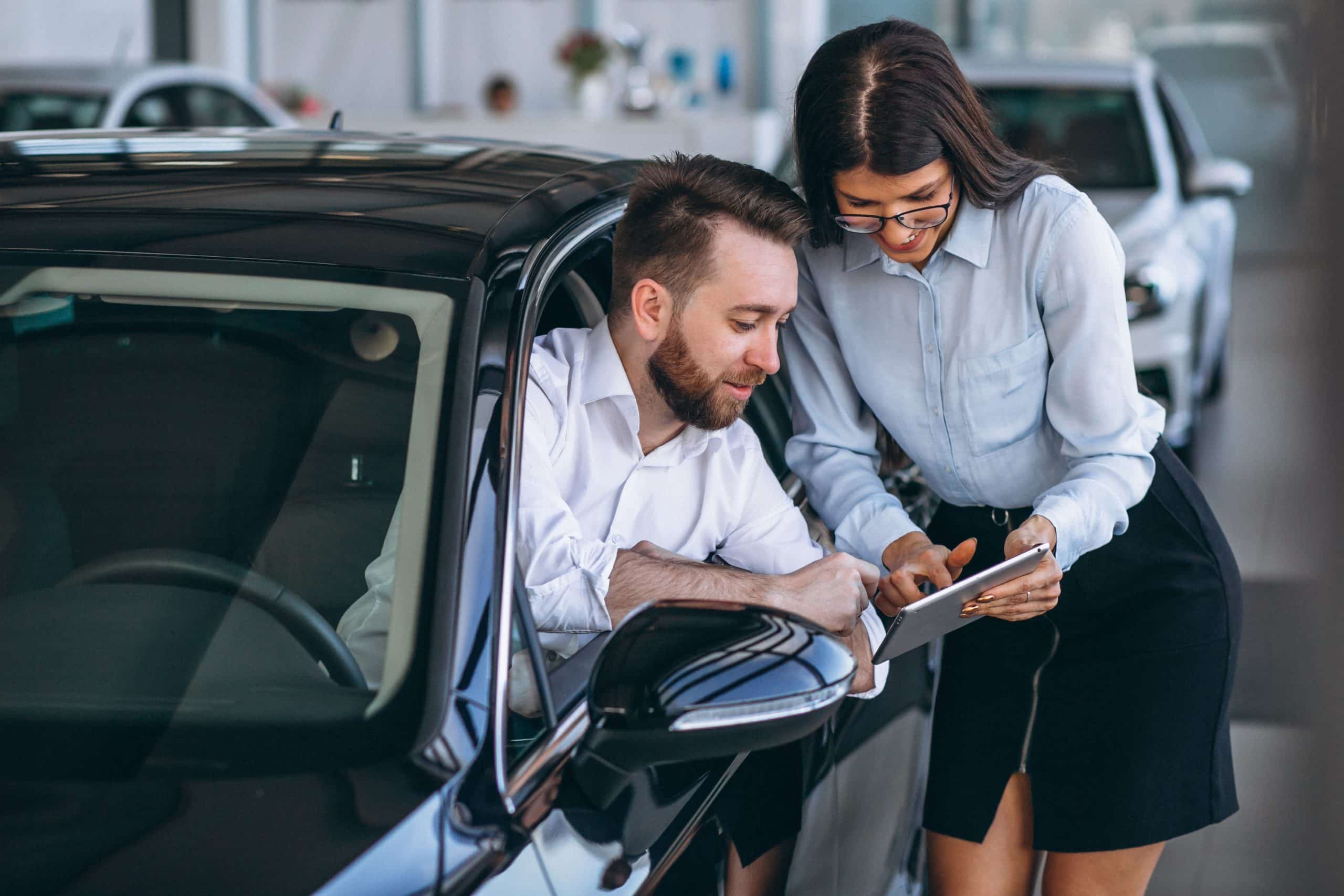 Car salesman and customer smiling while reviewing vehicle information on a tablet, with the man sitting inside a black car at the dealership, indicating a positive car buying experience.