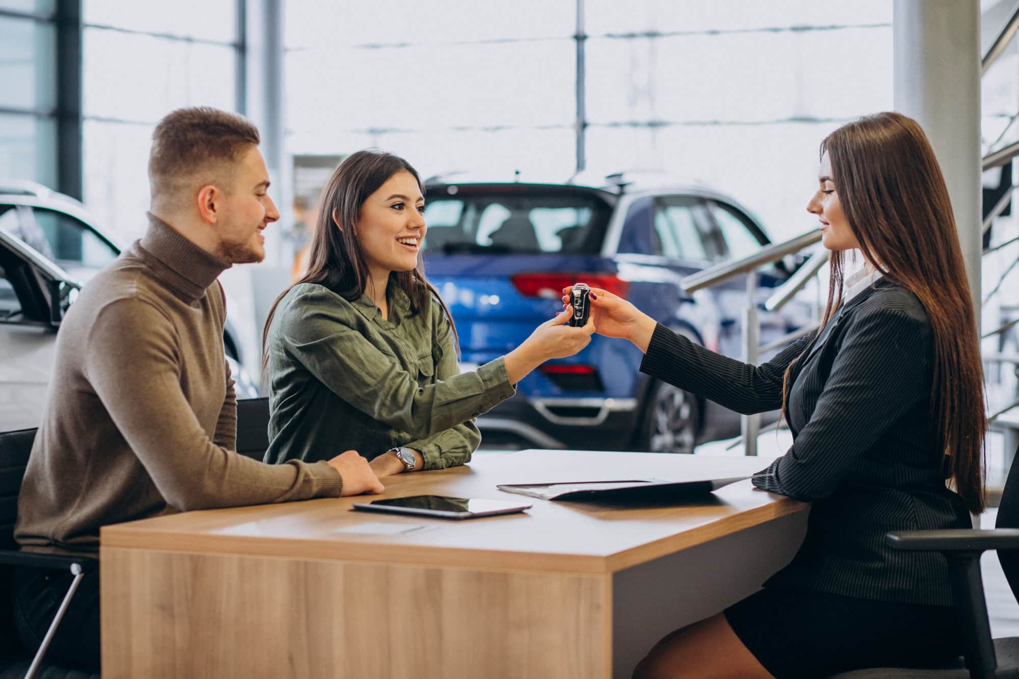 Young couple smiling as a car saleswoman hands over the keys at a dealership, representing the successful completion of a vehicle purchase.
