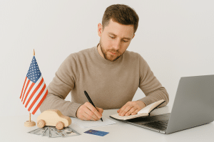 A man in his early thirties sitting at a white table, focused on paperwork related to vehicle financing. He is surrounded by a laptop, a toy car on top of dollar bills, a credit card, and a small American flag, in a minimalistic and well-lit setting.