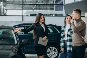 A car saleswoman showing a black vehicle to a smiling couple inside a modern car dealership. The woman is holding a tablet and gesturing toward the car, while the couple looks interested and engaged.
