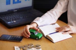 Close-up of a person at a desk holding a small green toy car while reviewing a planner. A smartphone with a calculator app open, coins, and a laptop displaying financial charts are also visible on the table. The image illustrates the process of budgeting and reflects concerns about inflation on vehicle financing, emphasizing how rising costs impact car affordability and personal financial planning.