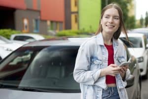 Young woman standing in front of a car while holding her smartphone, smiling and looking to the side. She is casually dressed, suggesting a relaxed and modern lifestyle. The image illustrates the concept of car finance for university students, highlighting the growing accessibility of vehicle ownership through tailored financial solutions for young adults pursuing higher education.