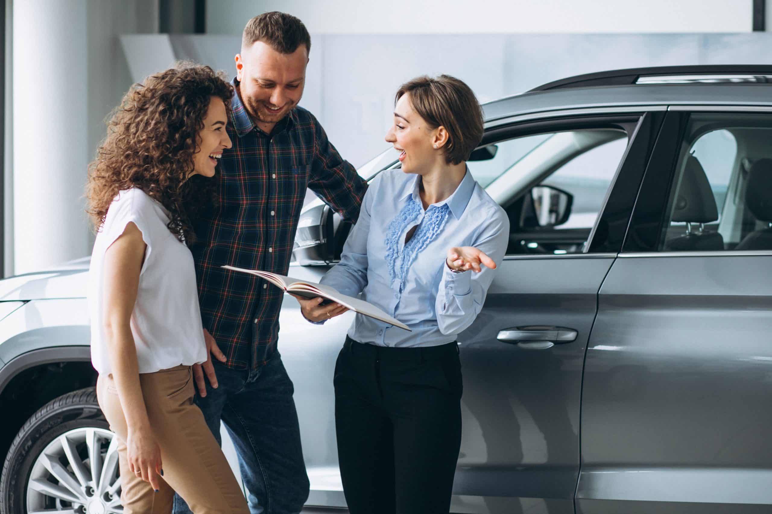A smiling couple is talking with a sales representative at a car dealership while reviewing information in a brochure. They appear excited and engaged in the discussion, standing next to a gray vehicle. The image represents the process of exploring options to get a car loan with no credit history, highlighting the guidance and support available for first-time buyers.