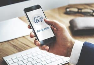 A person in a business suit holds a smartphone displaying an automobile icon, suggesting the use of mobile applications in the automotive sector. The setting includes a keyboard and wallet on a desk, emphasizing a professional environment where Artificial Intelligence may be enhancing digital car services and intelligent finance management.