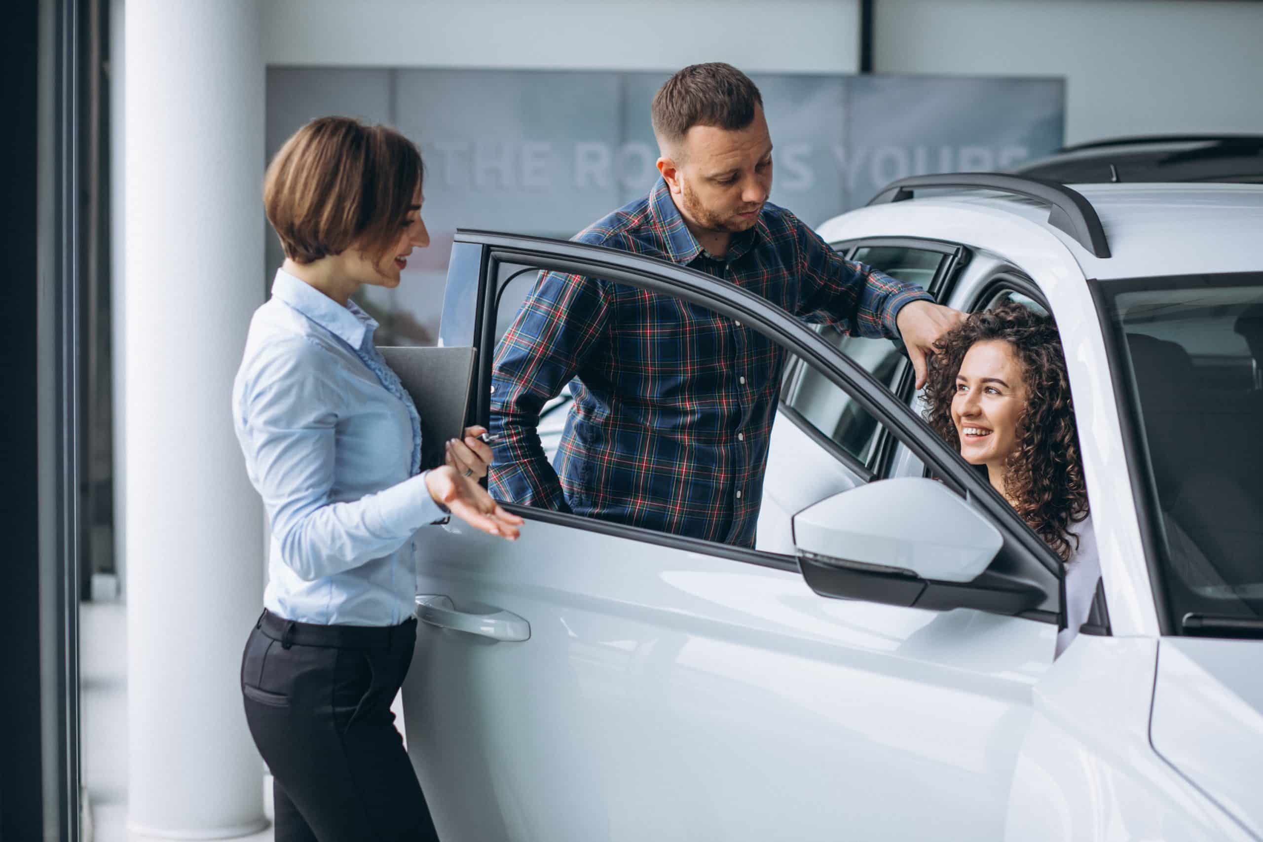 Smiling couple speaking with a saleswoman at a dealership while exploring a white SUV, illustrating the customer experience in car financing.