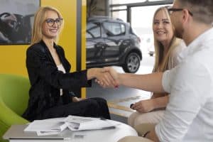 A smiling sales representative shakes hands with a couple at a car dealership, symbolizing a successful agreement on financing certified pre-owned (CPO) vehicles.