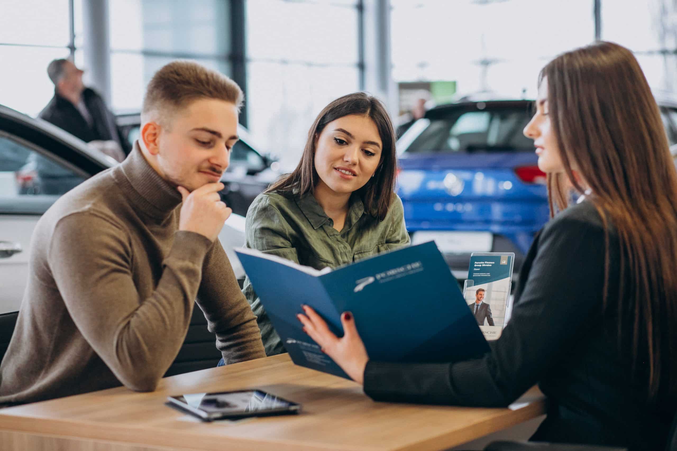 A couple sits at a desk in a car dealership, attentively reviewing financing documents presented by a female sales consultant. They are surrounded by vehicles, with a blue car visible in the background. The setting suggests a professional discussion about purchasing options, reflecting accessible vehicle financing for gig economy workers seeking flexible ownership solutions.