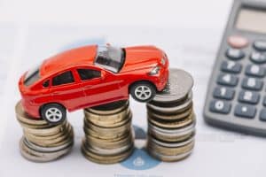 A small red toy car is positioned atop neatly stacked piles of coins, symbolizing automotive investment. A calculator and financial documents are visible in the background, representing loan calculations and affordability analysis. This visual concept highlights the appeal of Zero Percent APR offers in car financing promotions.
