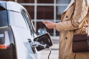A person wearing a beige trench coat is seen holding a smartphone while an electric car charges at a public charging station. The charging cable is securely plugged into the vehicle, emphasizing eco-friendly mobility. This scene illustrates the growing everyday use and convenience of electric vehicles in urban environments.