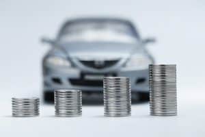 Stacks of coins increasing in height in front of a blurred car, symbolizing the rising impact of interest rates on vehicle financing.