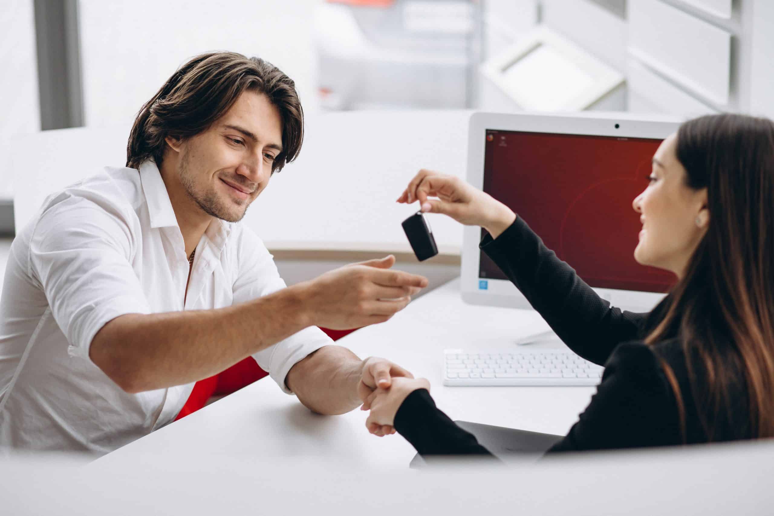 A smiling man in a white shirt receives car keys from a female dealership representative seated across the desk. A desktop computer is visible in the background, suggesting a professional setting. This scene represents a successful car purchase process, highlighting the convenience and efficiency of real-time credit approval on dealerships.