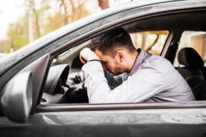 A frustrated man sits behind the wheel of a parked car with his head resting on the steering wheel, expressing stress or disappointment. The scene suggests financial strain or regret, often associated with issues like unexpected maintenance costs or vehicle devaluation over time.