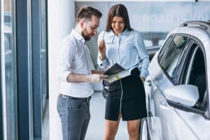 Man and woman standing next to a white SUV at a dealership, reviewing a brochure and discussing vehicle options, illustrating vehicle financing for small businesses.