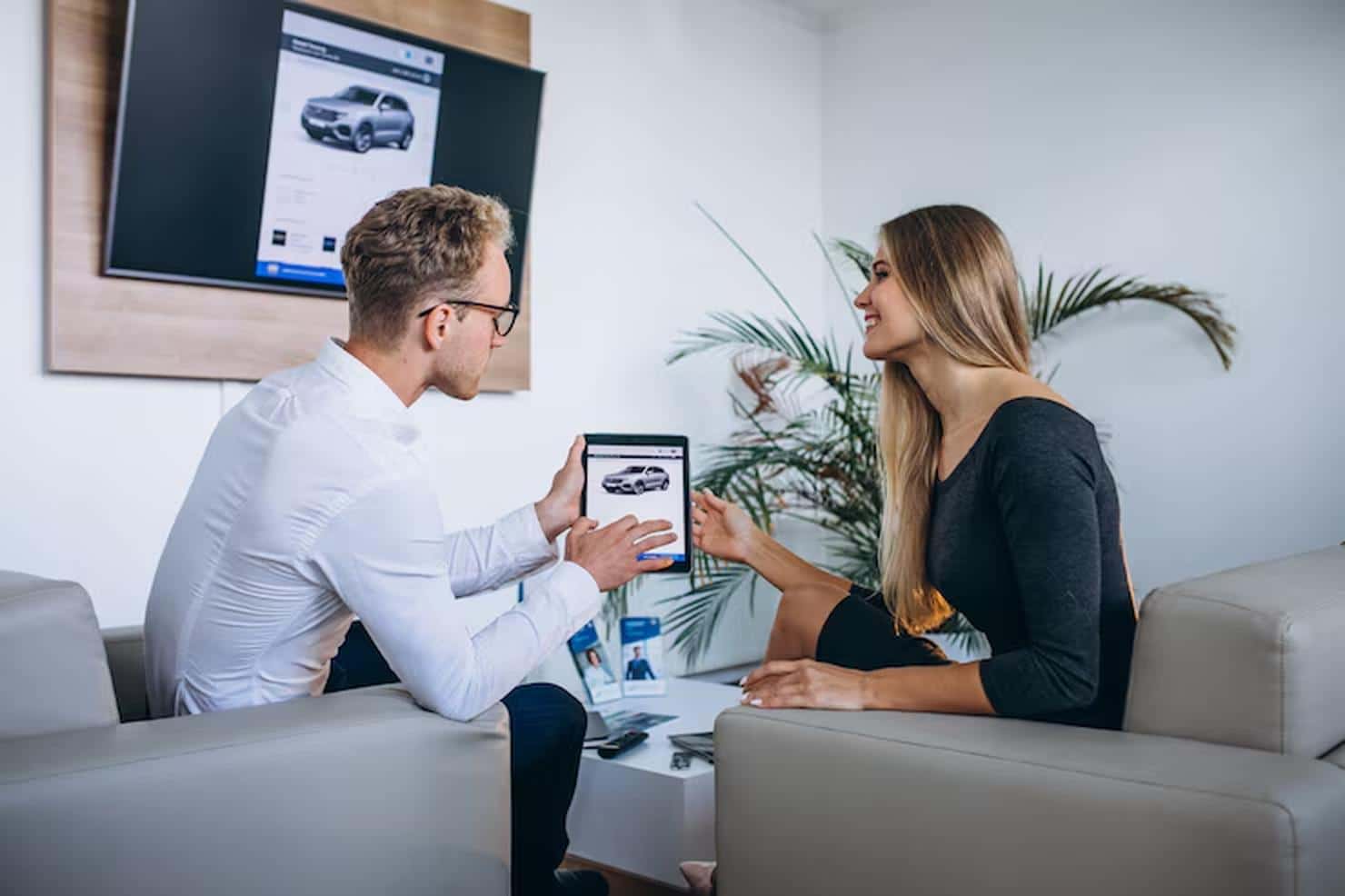 A man and a woman sitting in a modern car dealership lounge discussing Car Finance options while viewing a vehicle listing on a tablet, with the same car displayed on a screen in the background.