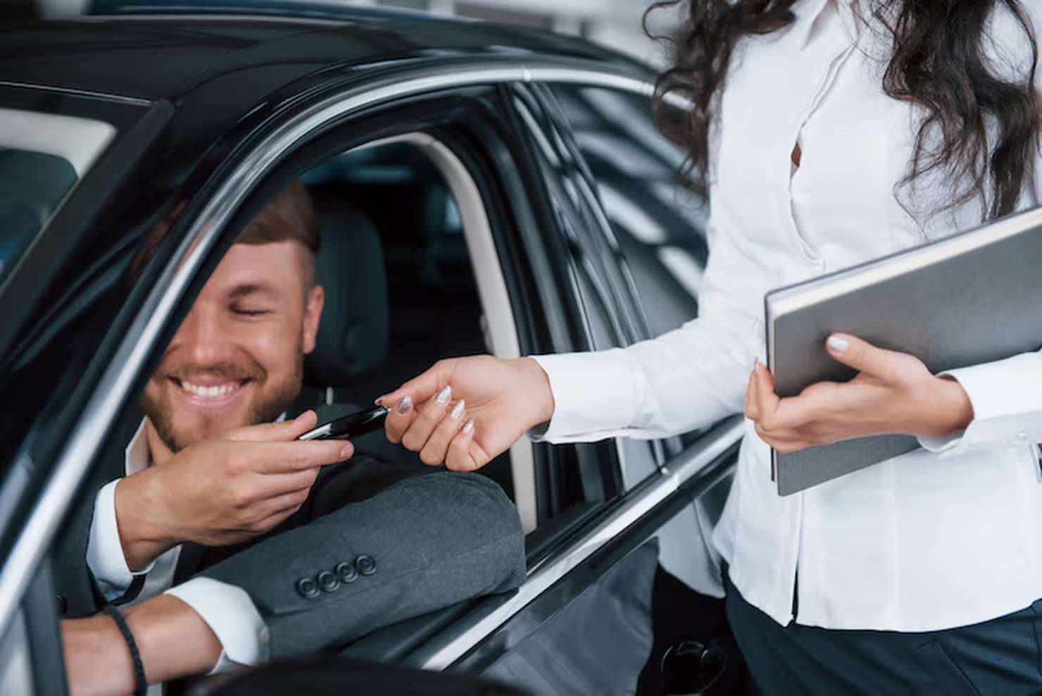 A smiling man sitting inside a car receives a key from a saleswoman holding a tablet, representing the successful completion of vehicle purchase through dealership financing programs.