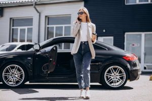 A stylish woman standing beside a sleek black car with the door open, holding a coffee cup and talking on the phone, representing confidence and the lifestyle associated with luxury vehicles.
