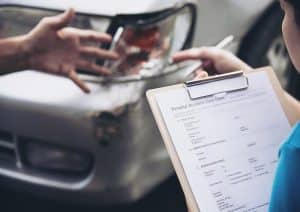 A person holding a clipboard with a personal accident claim form while inspecting damage to a car's front bumper, representing the types of incidents where no credit check car financing options may be considered for vehicle replacement or repair.