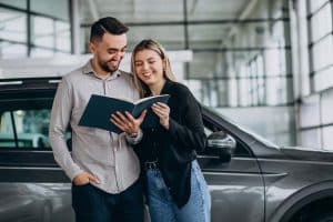 Smiling couple standing beside a car, reviewing a document or contract together, representing the decision-making process involved in payment car finance and vehicle acquisition.