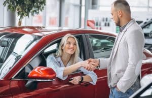 A smiling woman inside a red car shakes hands with a dealership representative, possibly finalizing a deal that includes options like extended warranty in vehicle financing.