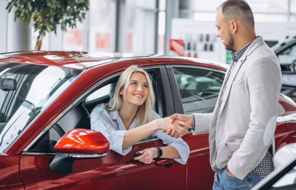 A smiling woman inside a red car shakes hands with a dealership representative, possibly finalizing a deal that includes options like extended warranty in vehicle financing.