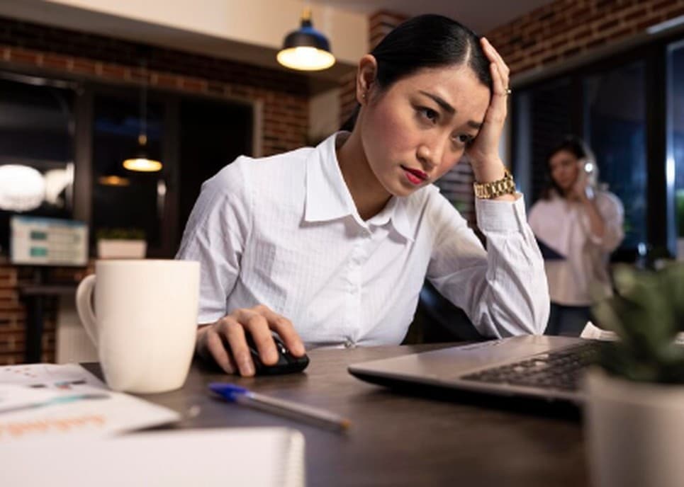A concerned woman sits at a desk in a dimly lit office, looking at her laptop with a hand on her forehead, symbolizing the stress of unexpected career transitions and the importance to prepare financially for professional changes or layoffs.