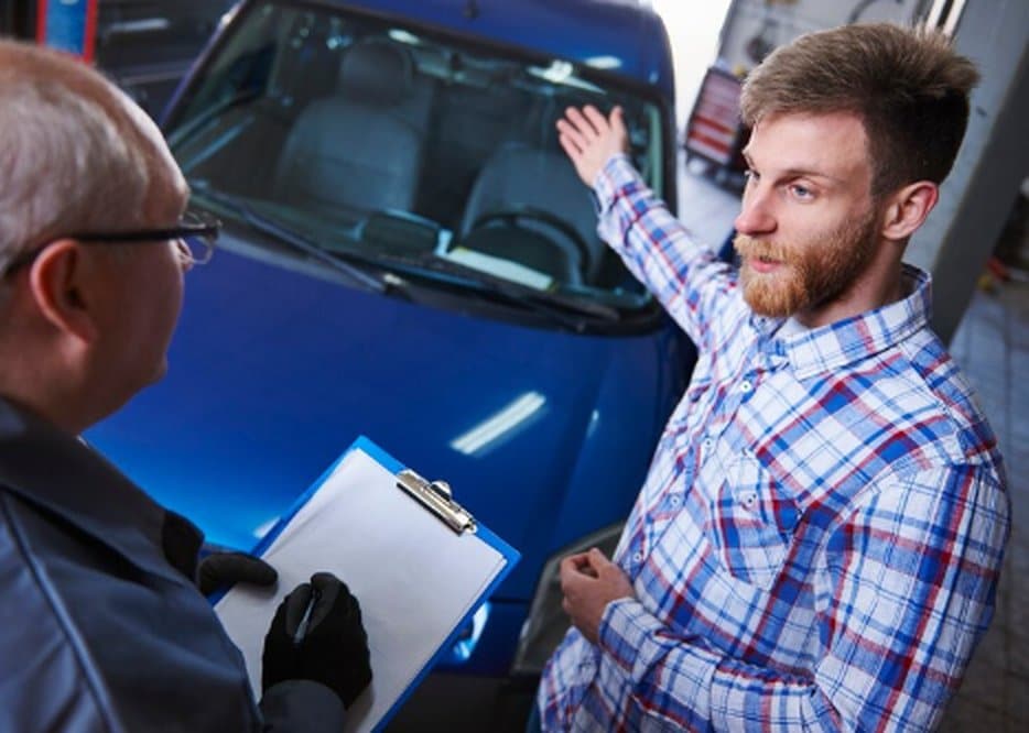 A man in a plaid shirt appears to be discussing the condition of a used car with a mechanic, likely during a pre-purchase inspection at an auto repair shop.