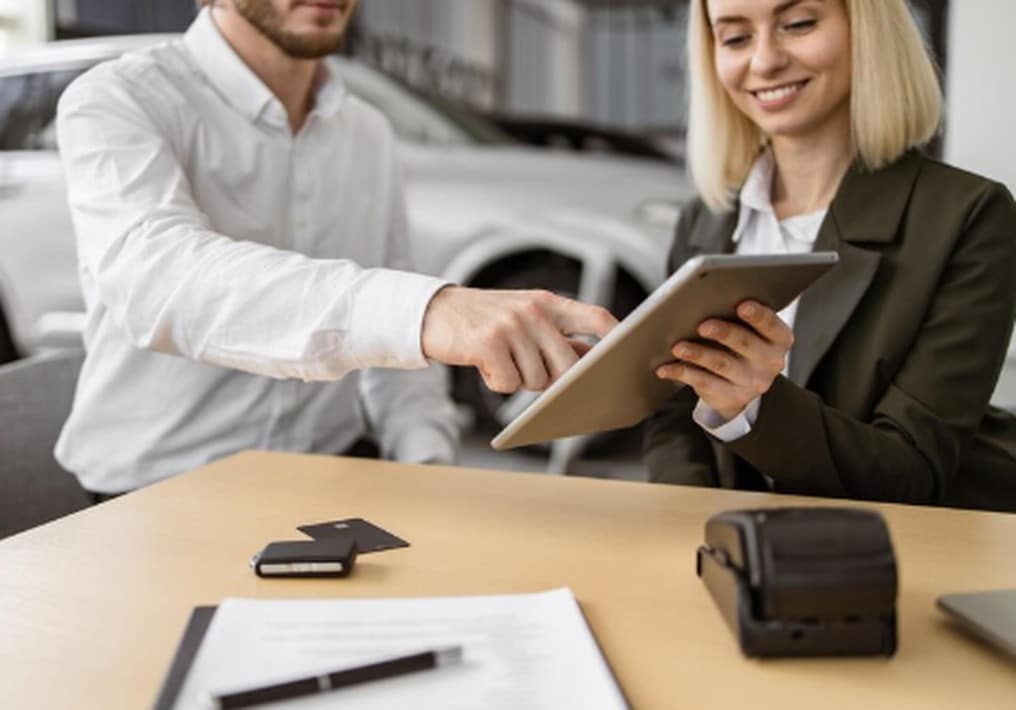 A smiling saleswoman shows a tablet to a customer at a car dealership, possibly reviewing digital documents or discussing car financing conditions before closing the deal.