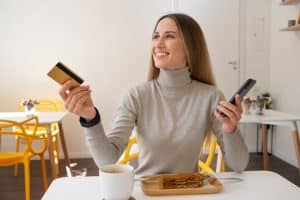 A young, smiling woman sitting at a cafeteria table, holding out her credit card to make a payment.