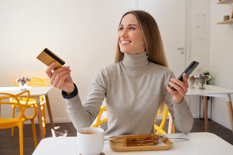 A young, smiling woman sitting at a cafeteria table, holding out her credit card to make a payment.