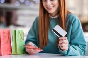 Smiling young woman holding a credit card and a smartphone while shopping, with colorful gift bags on the table, illustrating convenient and secure purchases using a credit card.