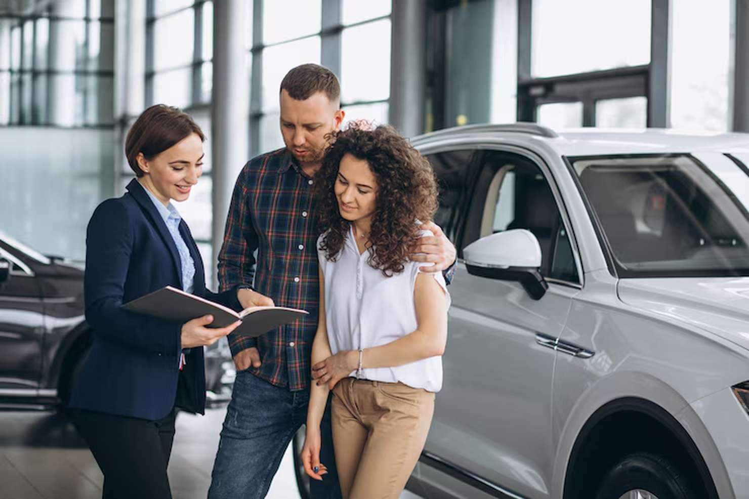 A smiling car saleswoman shows a couple a contract for car financing while standing next to a shiny silver SUV inside a modern dealership.