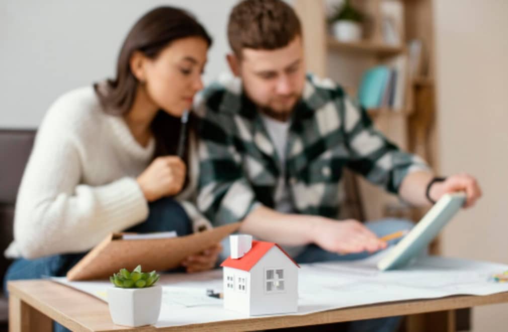 O ChatGPT disse: Couple reviewing documents and using a tablet at a table with a small model house in the foreground, symbolizing the analysis and signing of a financing contract for purchasing property.