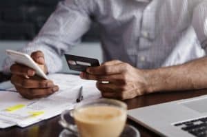 Man holding a credit card while checking his smartphone and reviewing bills on a desk, illustrating the challenges of managing revolving credit.