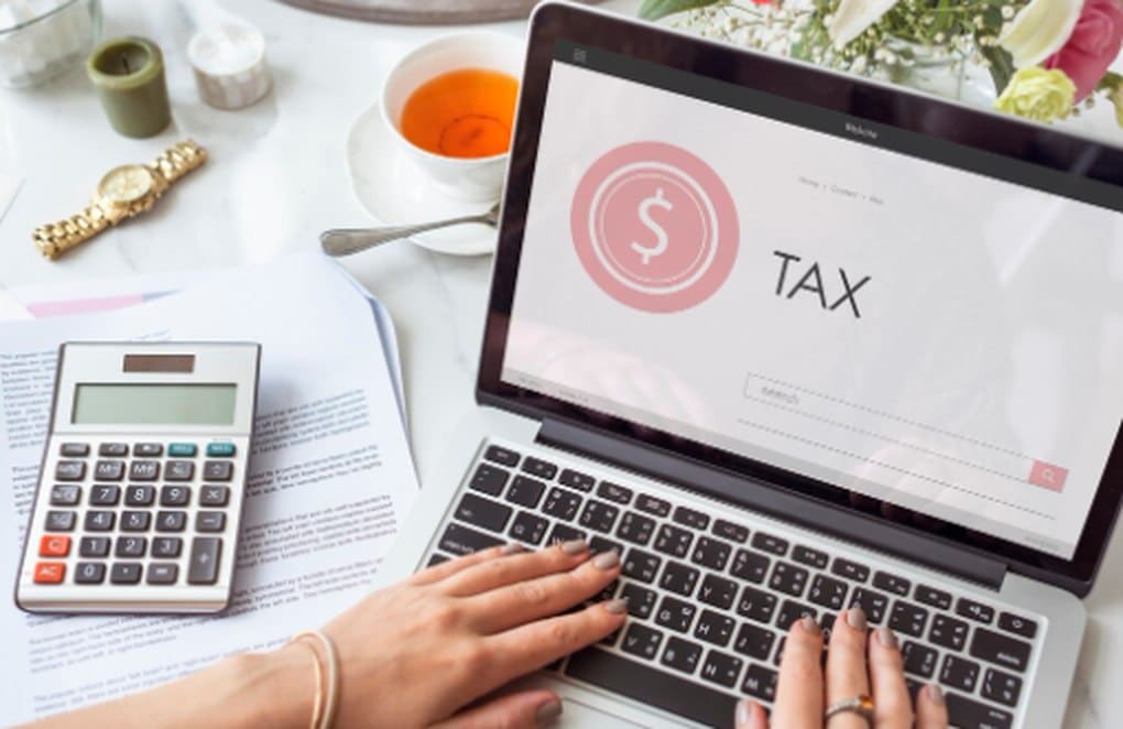 Person working on a laptop with an online tax application, calculating expenses and managing tax refunds with supporting documents and a calculator on the desk.