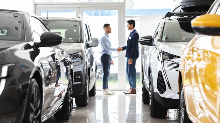 Two men shaking hands inside a car dealership, surrounded by new vehicles, symbolizing a business deal for commercial car leasing.