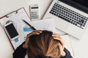 Person holding their head in frustration while sitting at a desk with financial documents, a laptop, and a calculator, illustrating stress and challenges in financial management.