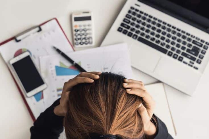 Person holding their head in frustration while sitting at a desk with financial documents, a laptop, and a calculator, illustrating stress and challenges in financial management.