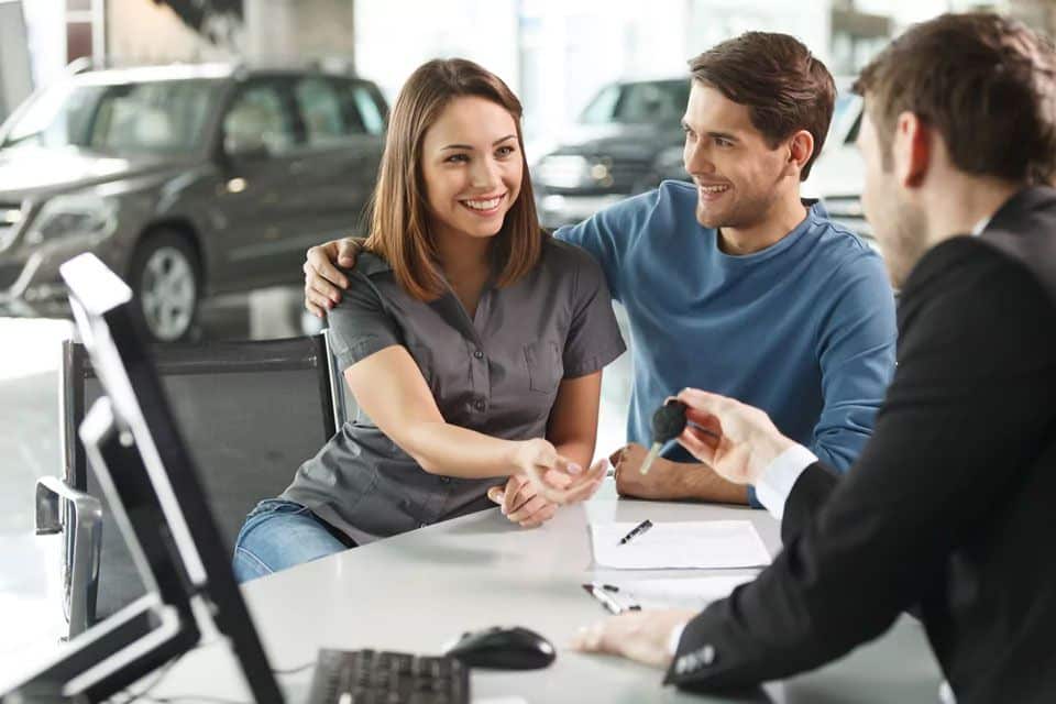 Smiling couple meeting with a car dealer at a showroom, signing documents and receiving car keys, symbolizing successful car financing and vehicle purchase approval.