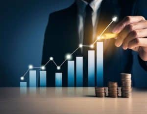 Businessperson drawing an upward arrow over a growing bar chart with stacked coins on a desk, representing financial growth, investment performance, and increased daily returns.